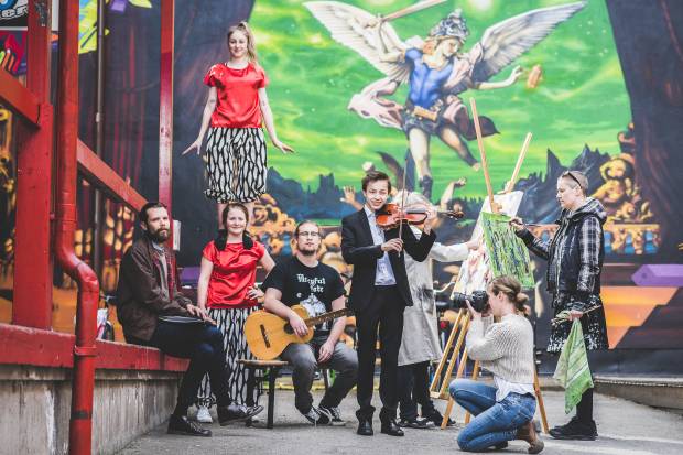 Flera personer står framför en graffitimålad vägg och utför olika typer av kulturella aktiviteter. Några gör akrobatiska konster, andra spelar instrument eller målar. 
